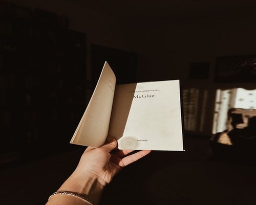A relaxed individual reading an open paperback book in a well-lit living room in Guadalajara with soft shadow patterns